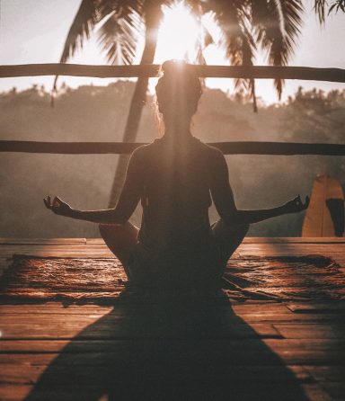 Silhouette of a woman meditating on a deck at sunrise, framed by palm trees and bathed in golden light.